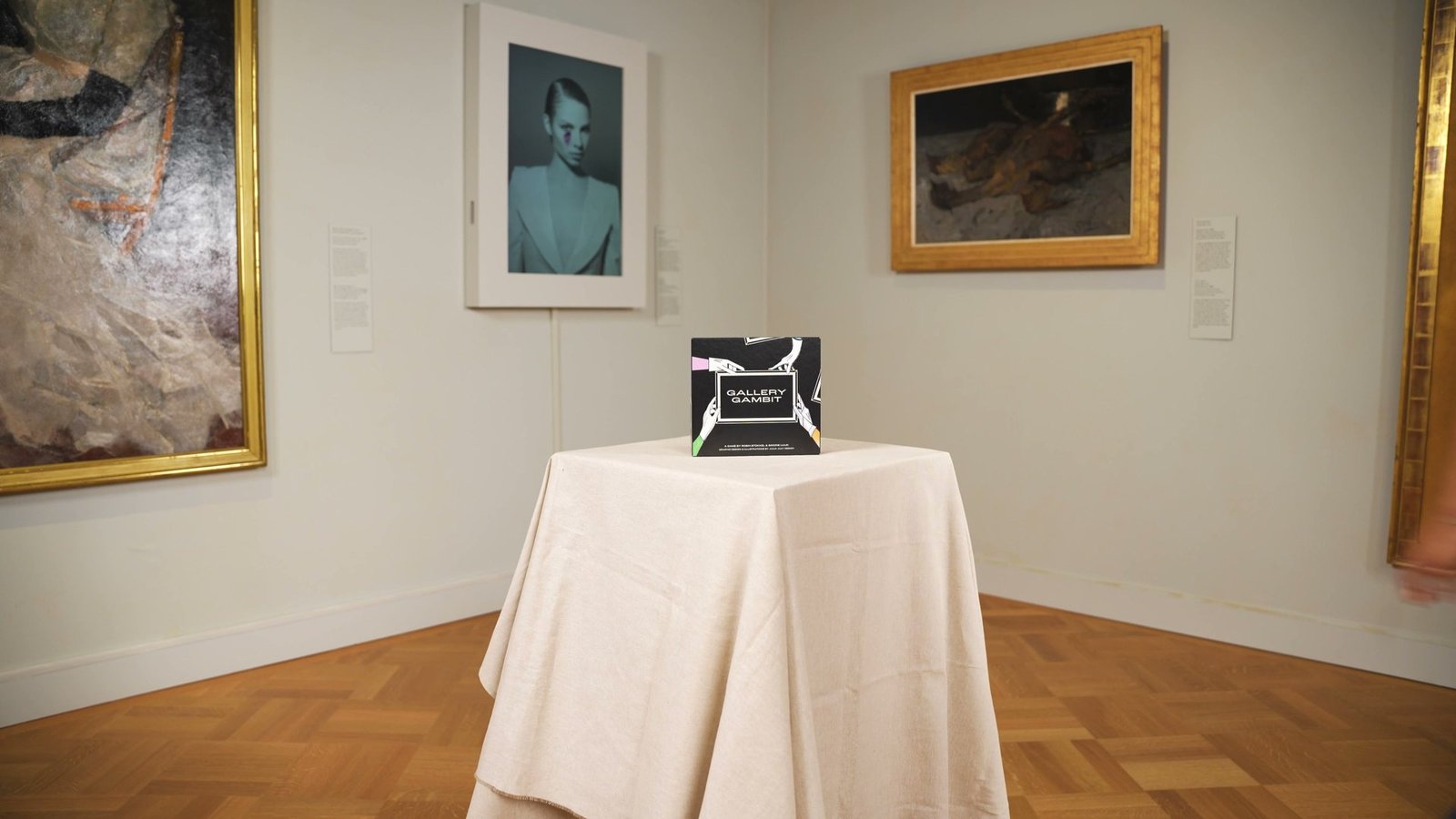 The Gallery Gambit box on a white plinth in a museum room, between framed paintings.