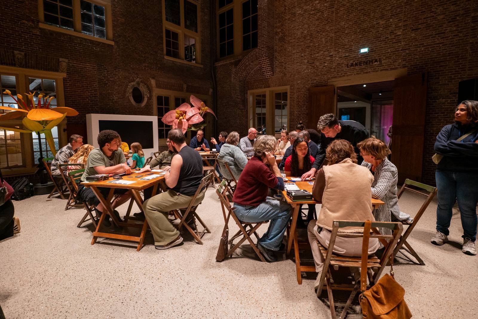 The Achterplaats of Museum De Lakenhal in the evening, full of people playing Gallery Gambit at wooden tables between giant paper flowers.