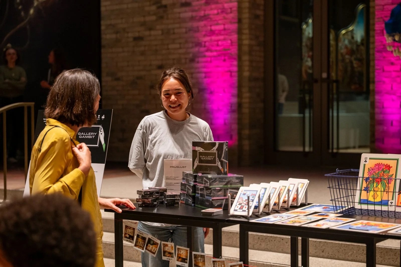 Julia July smiling behind the Gallery Gambit stand at Museum De Lakenhal, talking with a visitor in a yellow coat.
